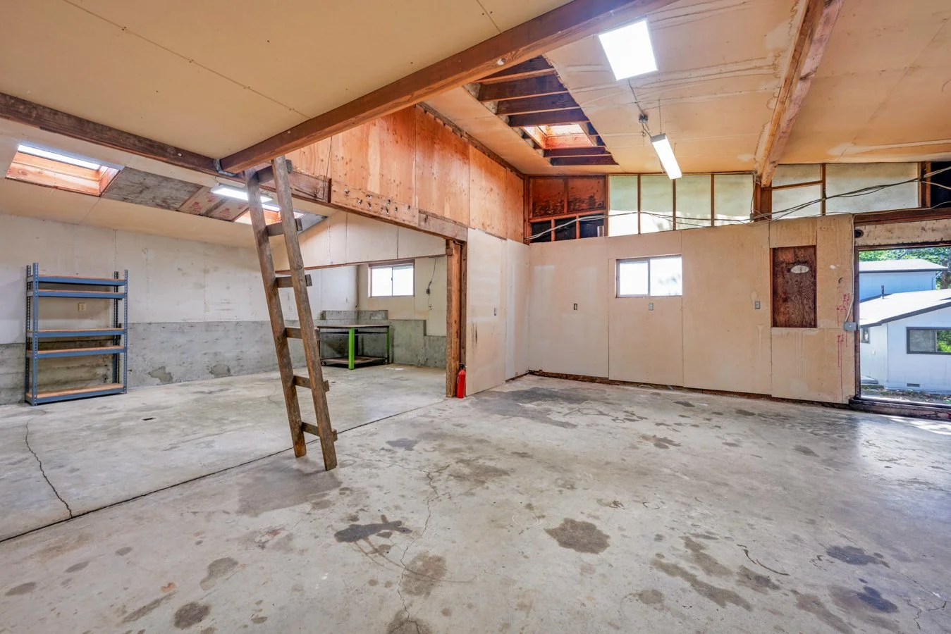 Interior of an unfinished garage or workshop with exposed concrete floors, wooden framing, and a ladder leaning against a wall. There's a shelf with empty racks on the left and small windows on the walls, with some natural light coming in.