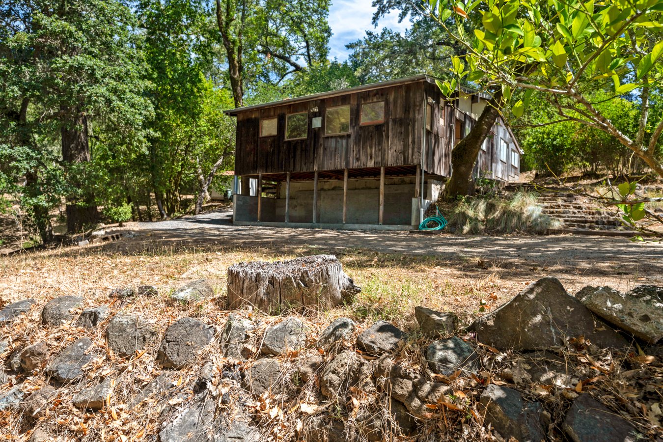A rustic wooden house elevated on stilts in a wooded area with trees and rocks surrounding it.