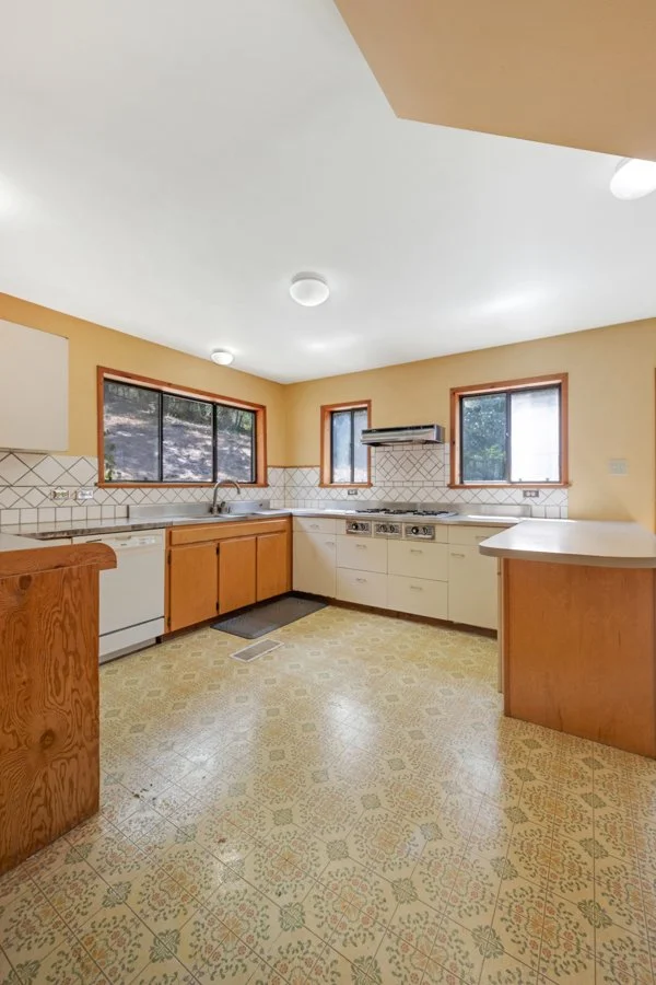 Empty kitchen with beige walls, wooden cabinets, tile backsplash, two large windows, and a gas stove. The floor has patterned linoleum.