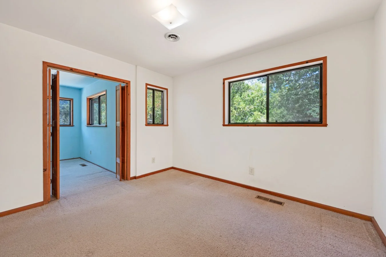 Empty room with white walls, beige carpet, multiple windows with wooden trim, and a doorway leading to another room with blue walls and windows.