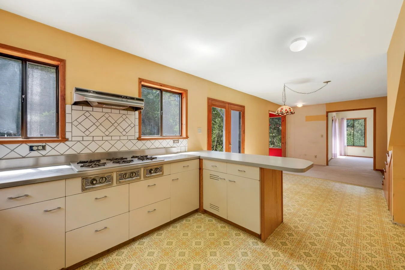 An empty kitchen with yellow walls, wooden window frames, white cabinets, a tiled backsplash, and a patterned yellowish floor. The kitchen has three windows, a sliding glass door, and a connected dining or living area with a hanging light fixture.
