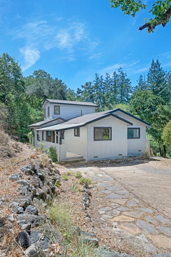 A two-story white house with black trim, surrounded by trees on a hillside, under a blue sky with some clouds.