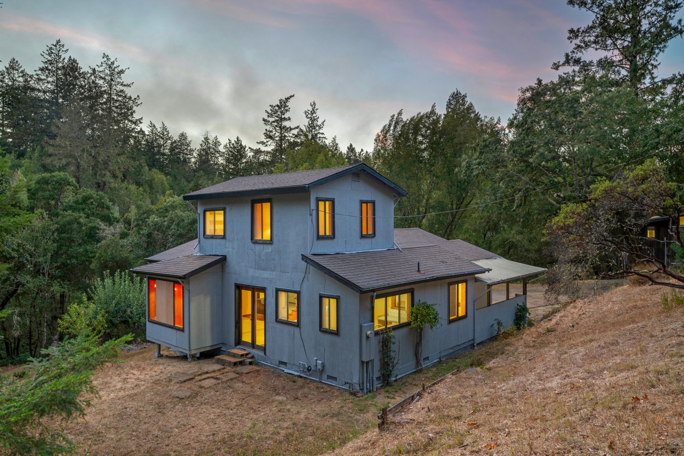 A two-story house with multiple windows illuminated from inside, situated on a hill surrounded by trees as it gets dark outside.