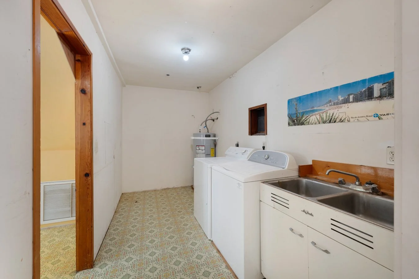 A laundry room with a white washing machine and dryer, a double stainless steel sink, and a small window on the wall. There is a water heater in the corner and a wall poster of a beach scene. The room has patterned linoleum flooring and plain white w