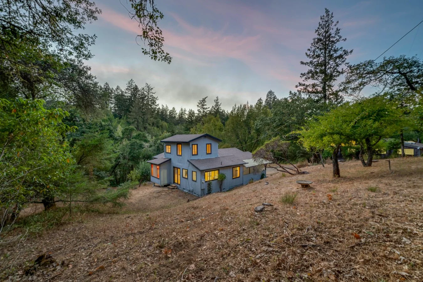 A modern blue house with yellow-lit windows situated on a slight incline amid a wooded area during dusk, with trees and a colorful sky in the background.