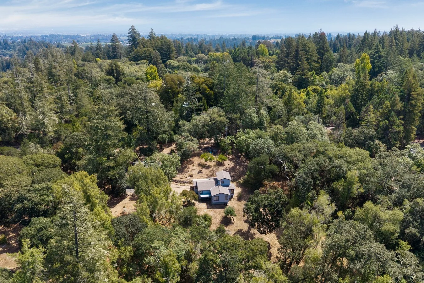 An aerial view of a house surrounded by dense green trees in a forested area with a distant cityscape in the background.