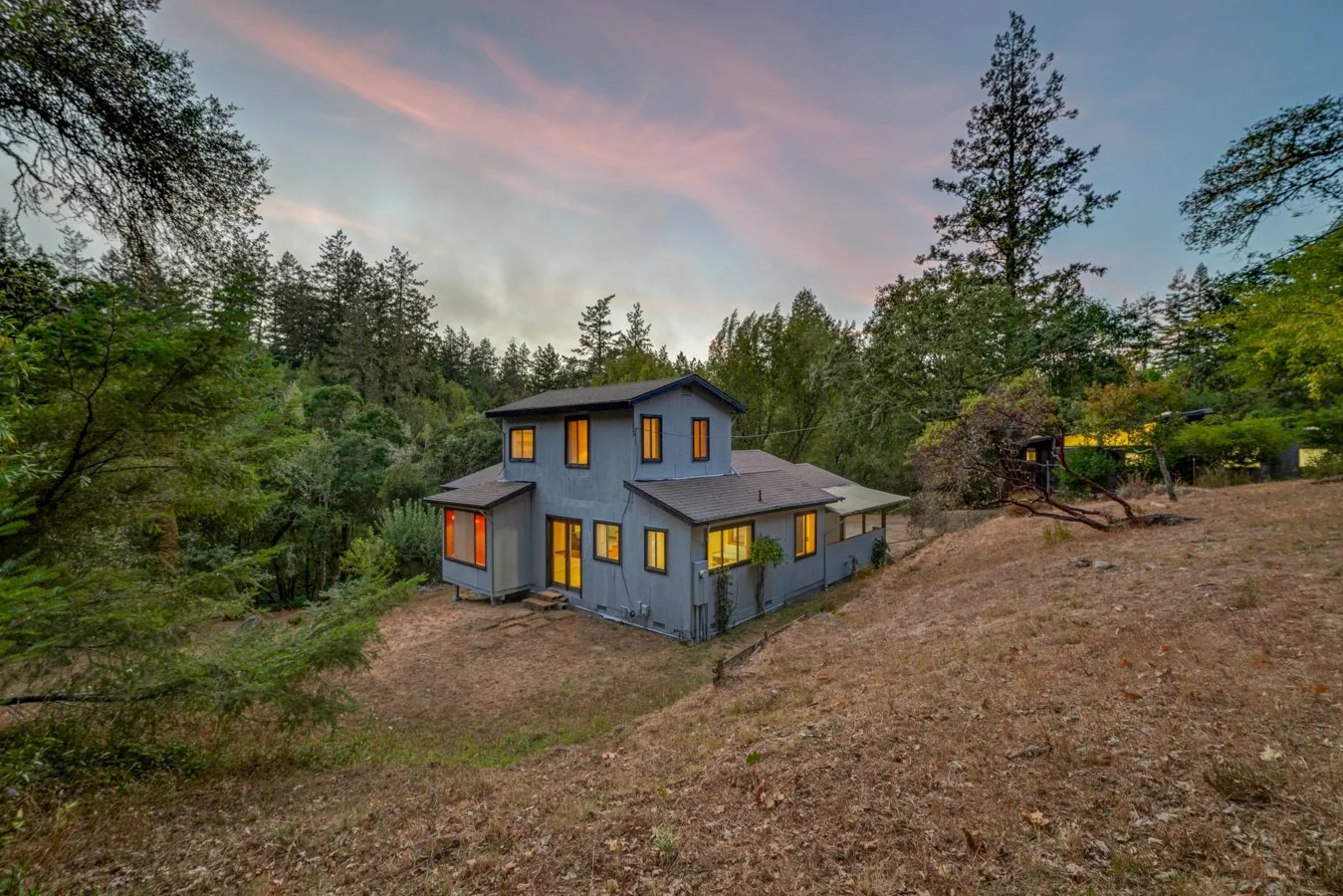 A two-story house with blue exterior walls and illuminated windows, situated on a sloped, dry grassy terrain surrounded by tall trees during sunset or dusk.