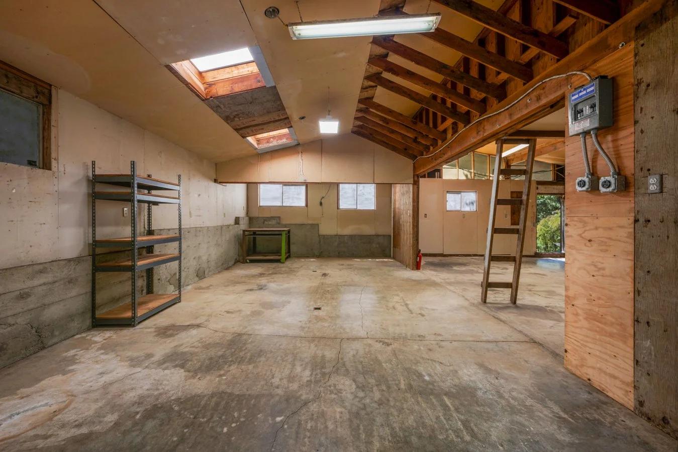 Empty garage or workshop space with concrete floor, wooden walls, and a sloped ceiling with skylights. There is a metal shelf on the left, a small green table at the back, and a ladder leading to a loft area.