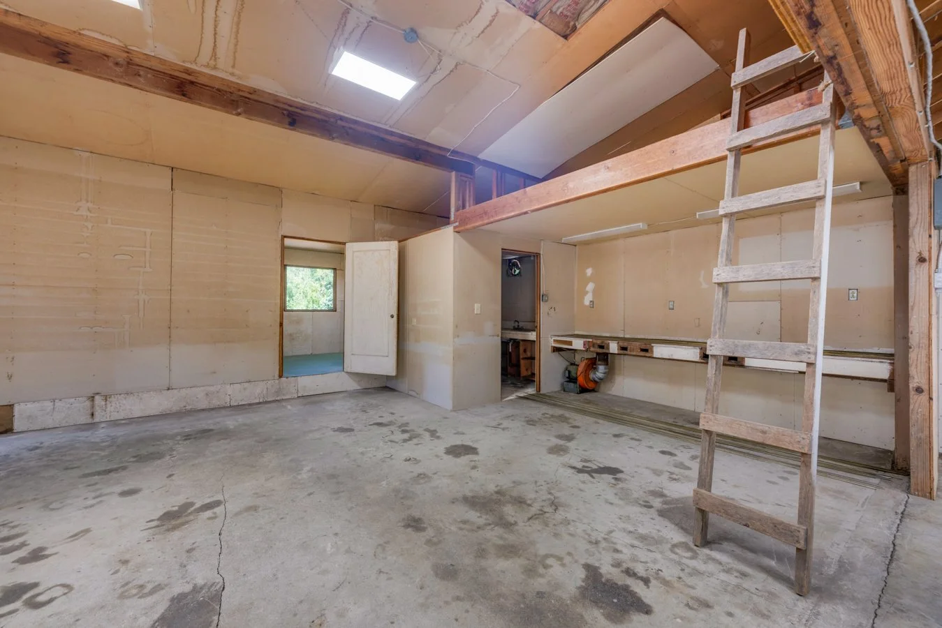 Empty unfinished room with drywall on the walls, a concrete floor, a wooden ladder, and a section of ceiling open showing wooden beams and insulation, possibly under renovation.