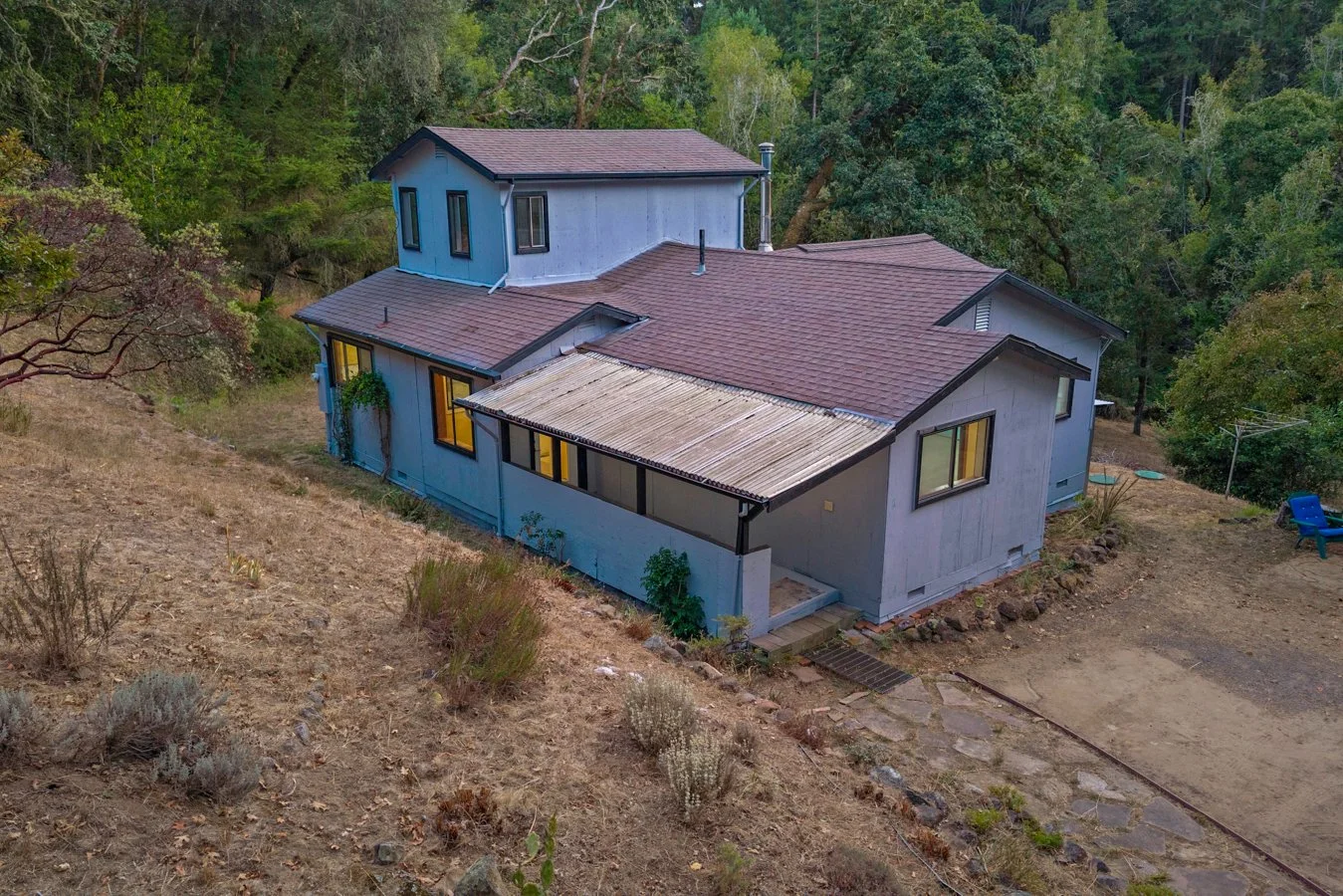 A house on a hillside surrounded by trees, with a dirt and stone pathway leading to the entrance, and a small patio area with a blue lawn chair