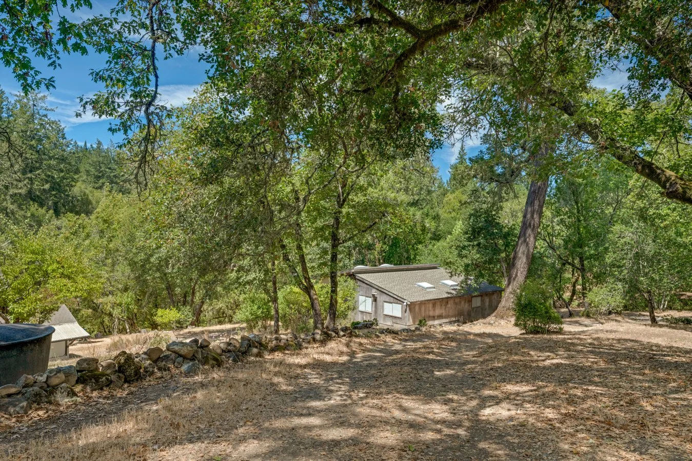 A house surrounded by trees in a wooded area with a dirt yard and a stone border.