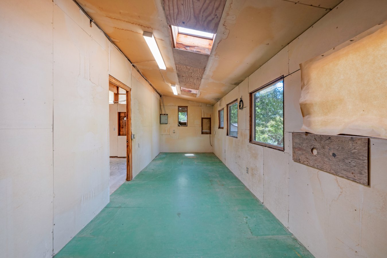 Room under construction with unfinished drywall, green floor, and windows showing trees outside.