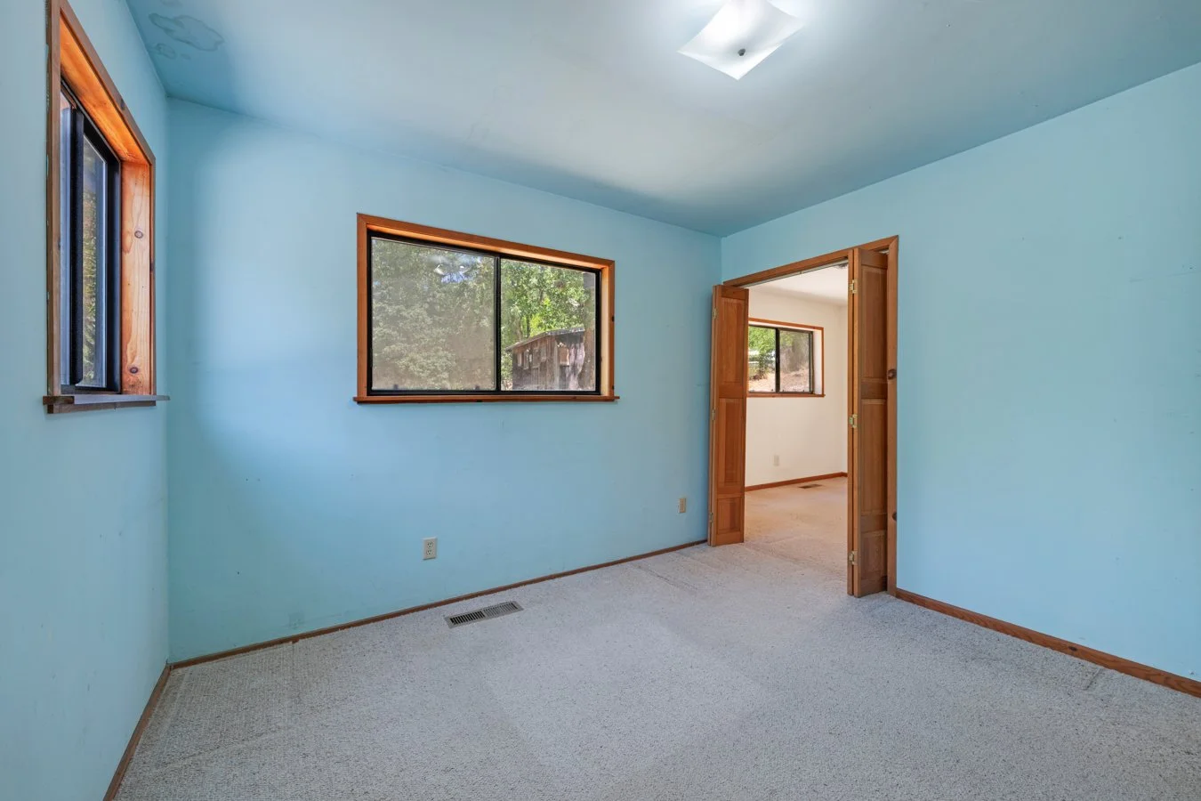 Empty room with blue walls, two windows, a ceiling light, beige carpet, and a wooden double door leading to another room with similar windows.