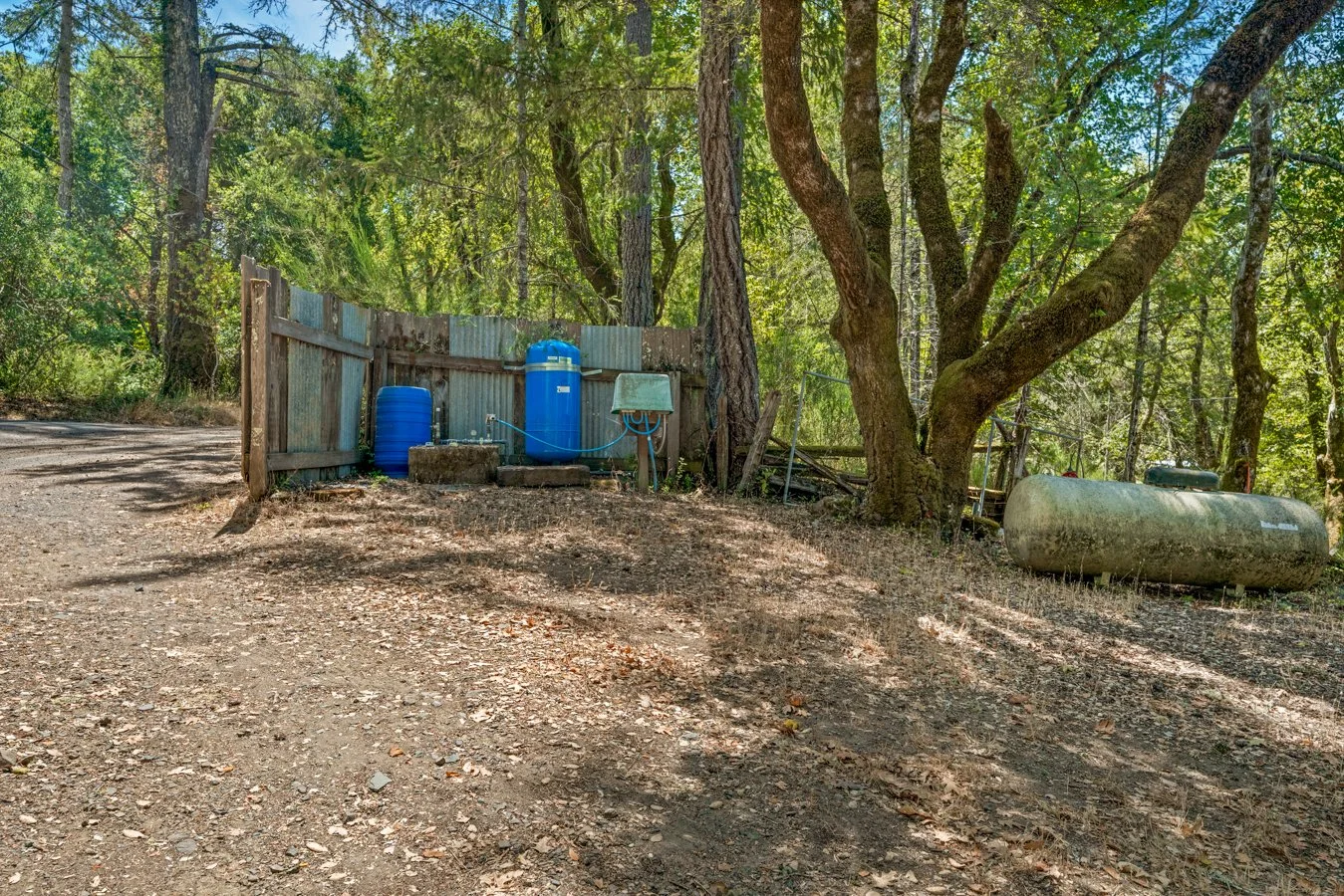 Outdoor scene with a dirt ground, a wooden and metal fence, and trees with green foliage. There are blue water storage tanks, a greenish propane tank, and various outdoor equipment near the fence.