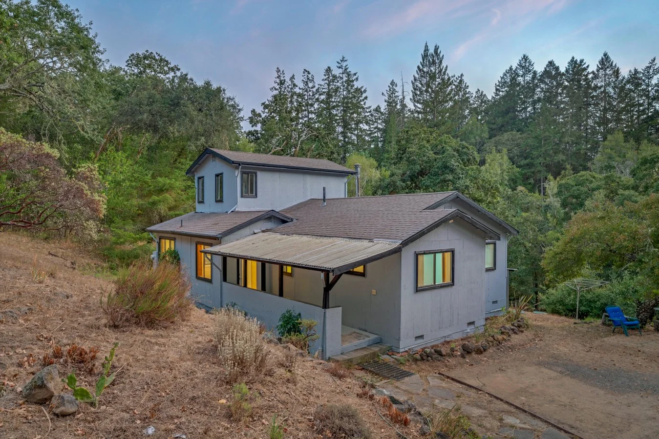 A two-story house with a smaller attached section, situated on a hillside surrounded by trees. The house has light-colored exterior walls, several windows with illuminated interiors, and a sloped roof. There is a dirt area in front, with rocks, dry p