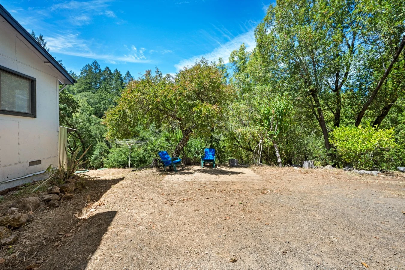 A backyard with a dirt ground, two blue Adirondack chairs, a small clothed clothesline, and lush green trees under a clear blue sky.