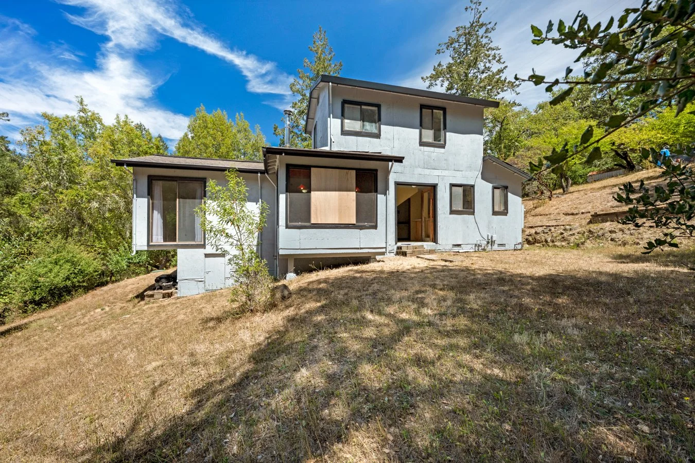 A modern, multi-story house with a gray exterior situated on a sloped dirt yard, surrounded by trees and under a partly cloudy blue sky.