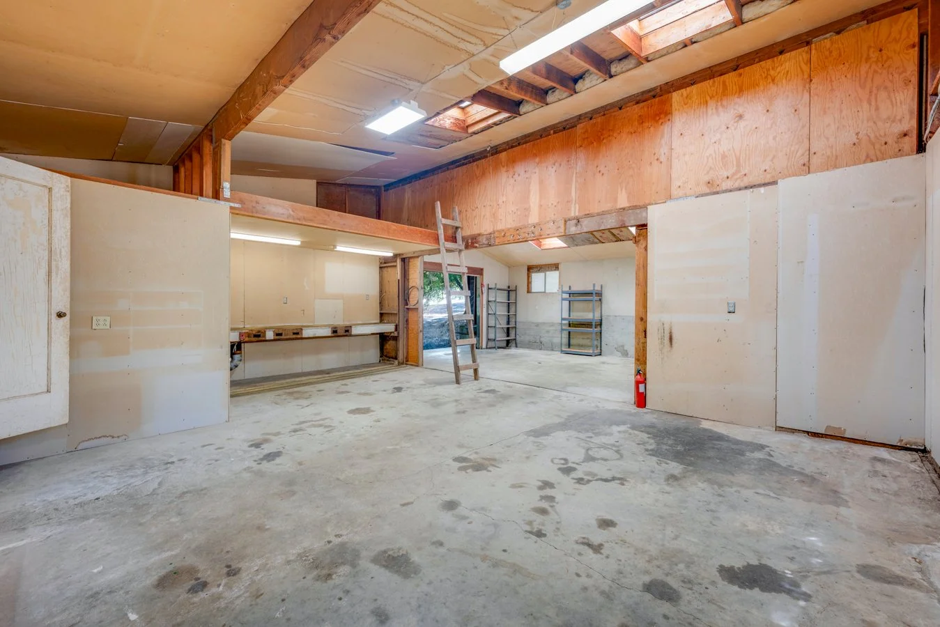 Empty garage with unfinished walls, exposed wooden beams, and a concrete floor; some shelves and a fire extinguisher visible.