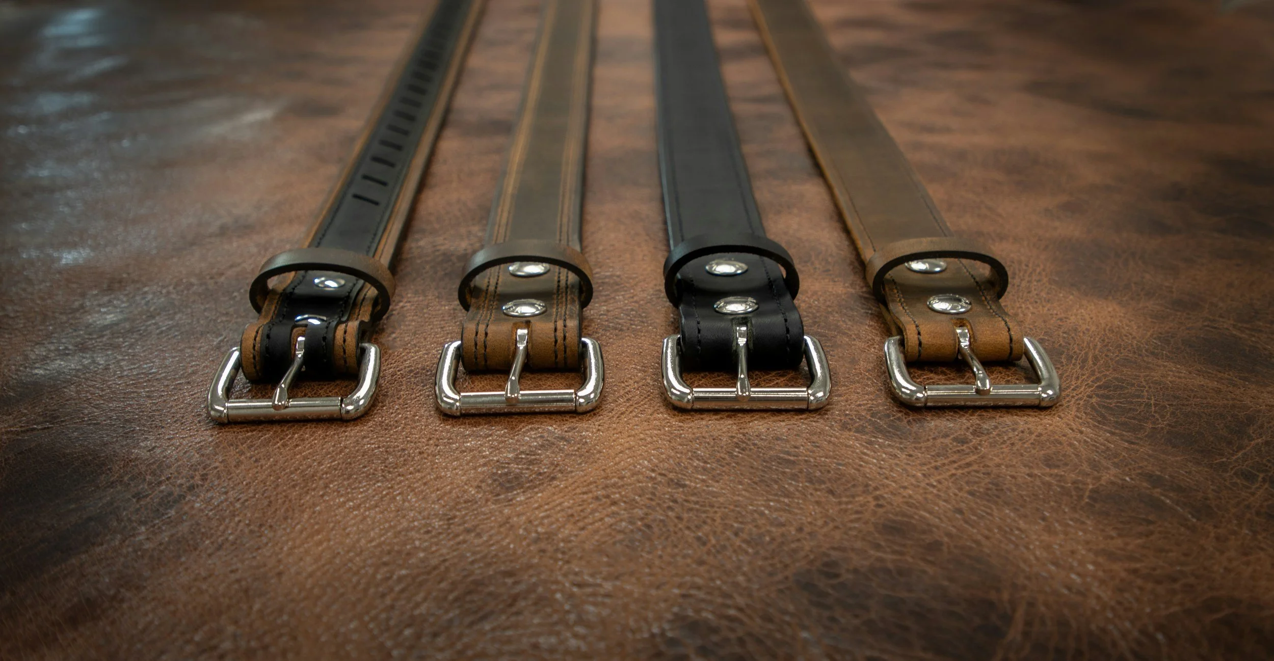Four leather belts with metal buckles laid out on a wooden surface.