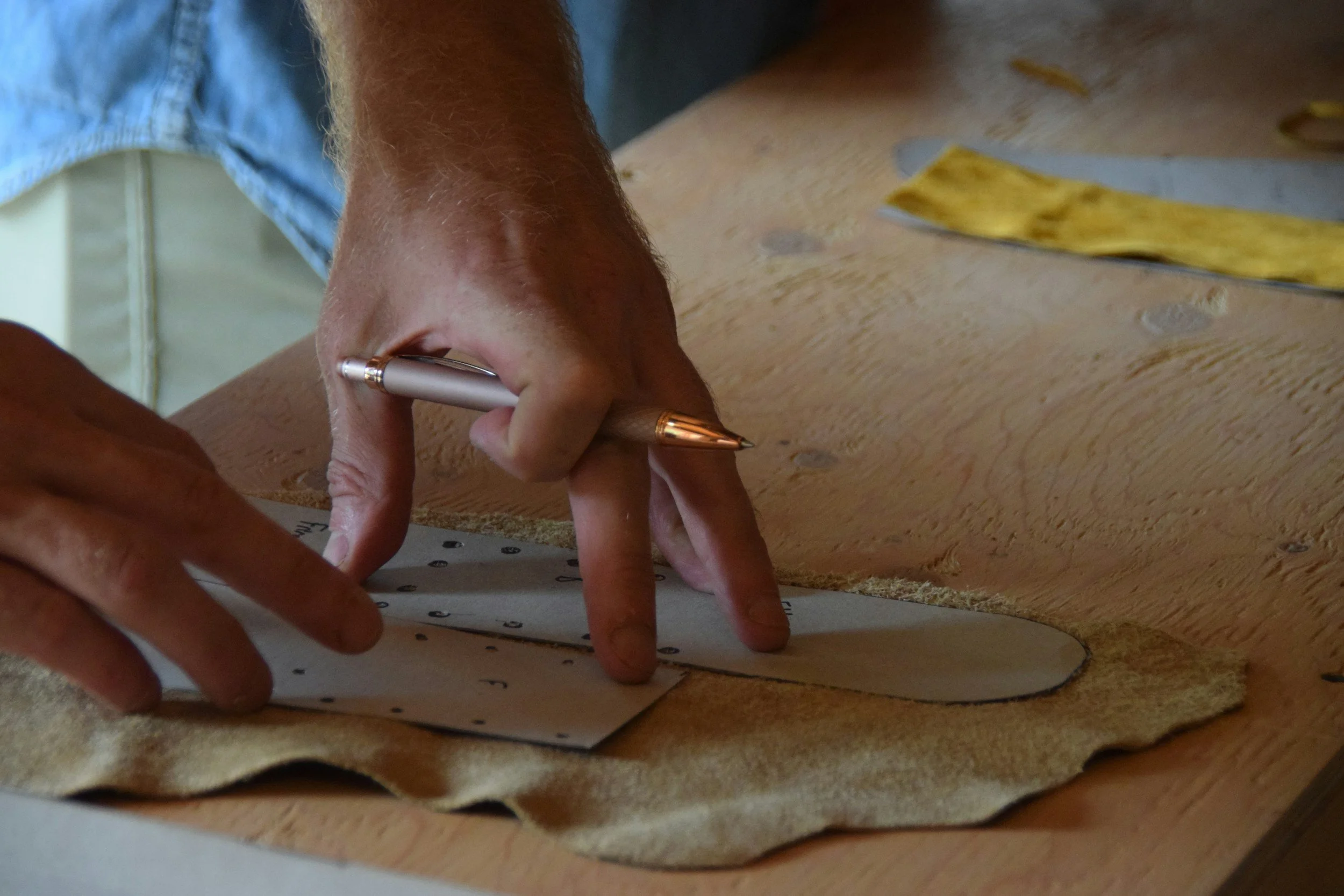 A person is using a silver pen to write on a piece of paper while working on a woodworking project. The project involves a piece of wood and a piece of sandpaper, indicating sanding or polishing.