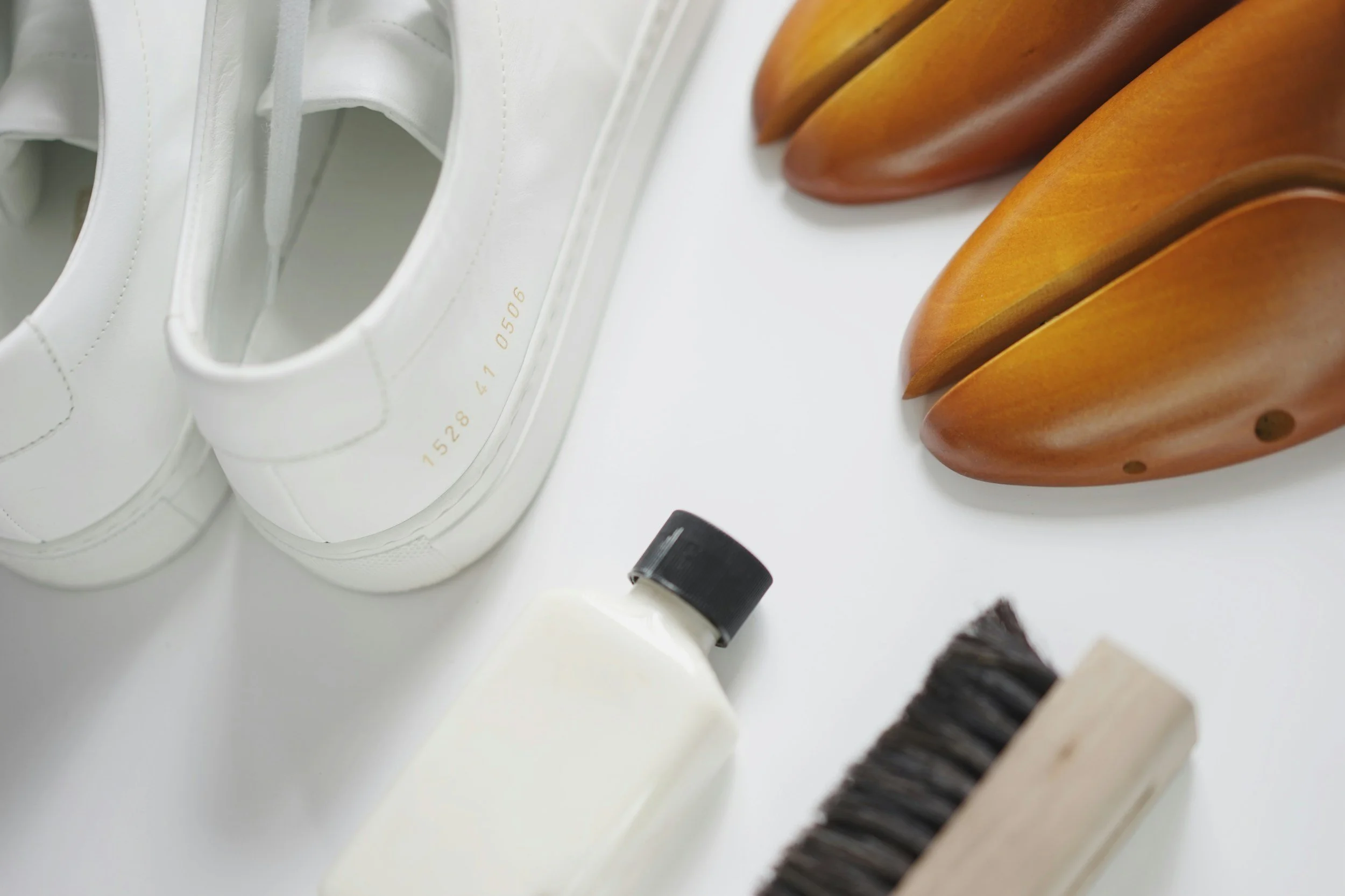Close-up of white sneakers, wooden shoe trees, and a small bottle of liquid in a white shoe storage or display area.