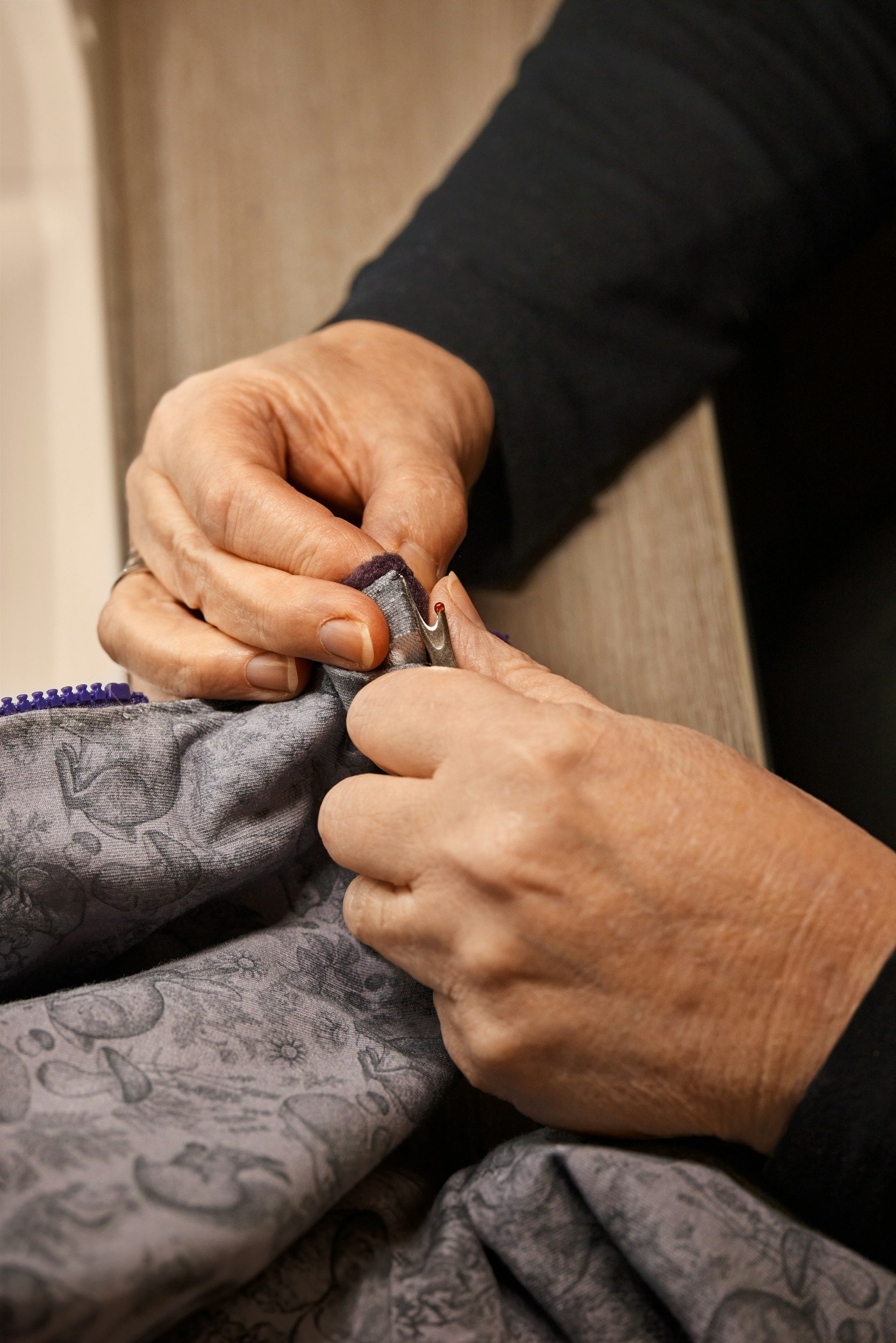 Close-up of two adult hands holding and possibly sewing or repairing fabric together. The hands are aged, and the fabric appears to be a gray patterned material.