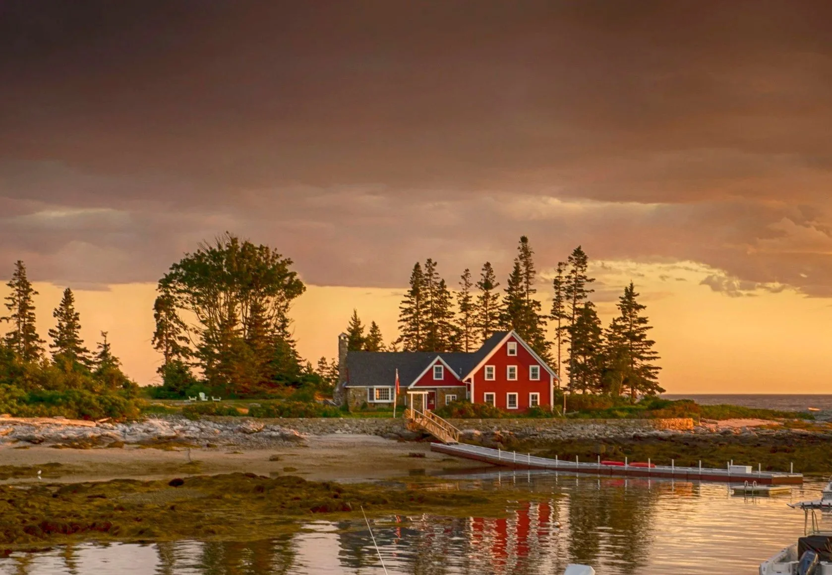 A red house on a shoreline during sunset with trees in the background and a dock extending into the water