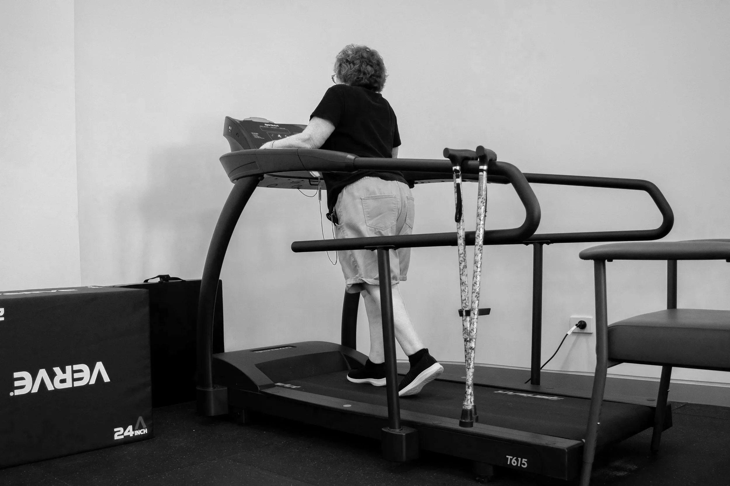 A woman with curly hair and glasses walking on a treadmill in a gym or fitness center. She is wearing a black shirt, light-colored shorts, and black athletic shoes. The treadmill displays the brand name "Vevor" and a size indicator "24 inch".
