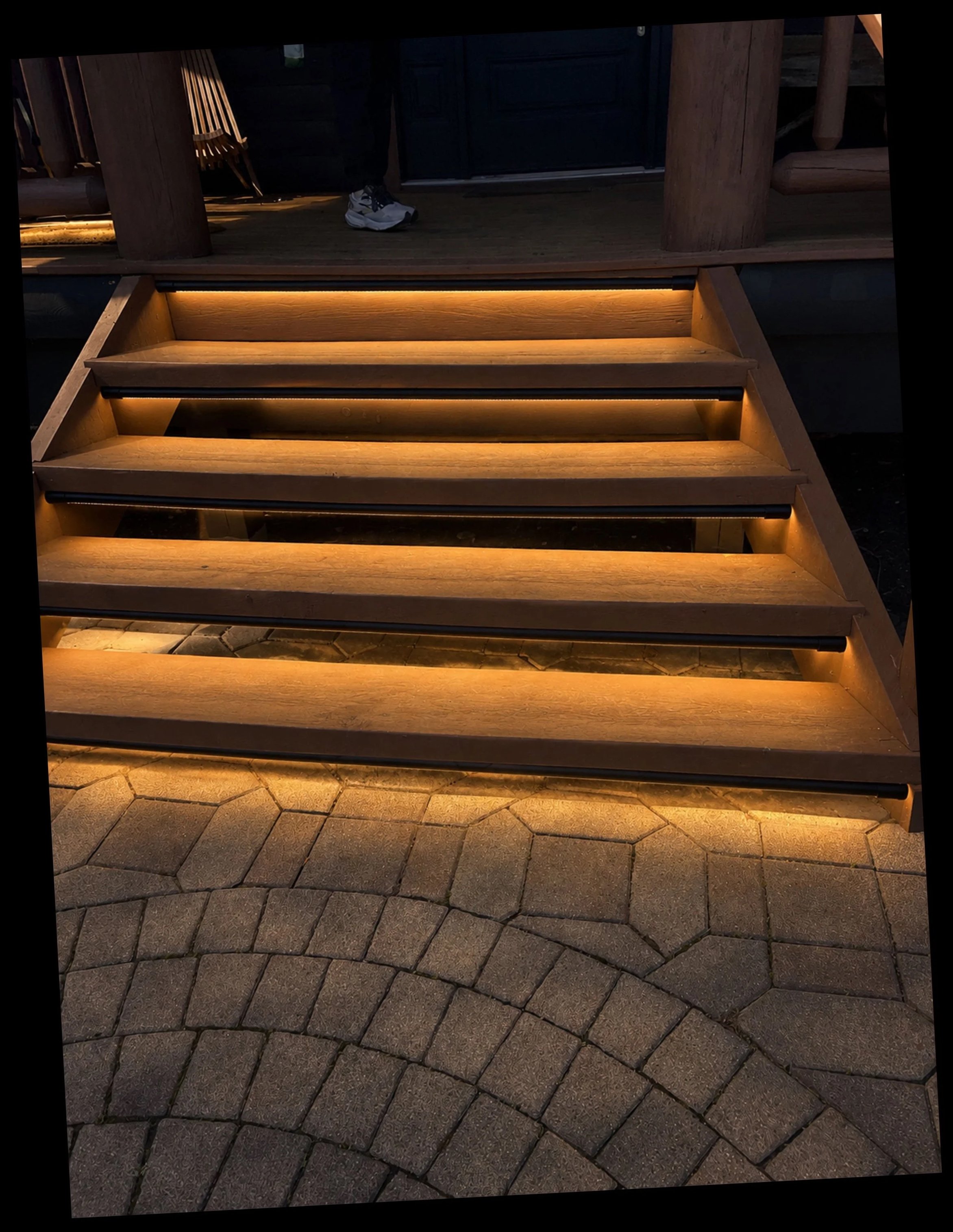 Wooden stairs with illuminated treads leading from a stone paved area to an outdoor deck. Part of a person's lower body and sneakers are visible at the top of the stairs.
