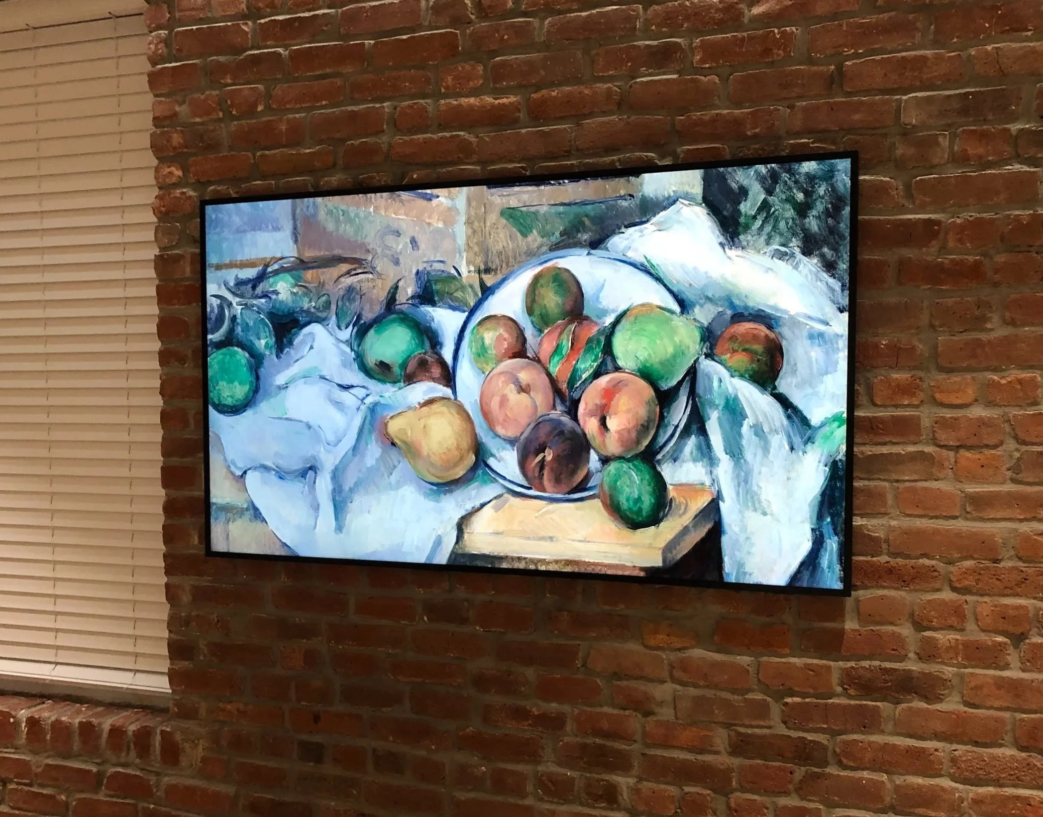 A painting of a bowl of assorted fruits on a table, hung on a brick wall.
