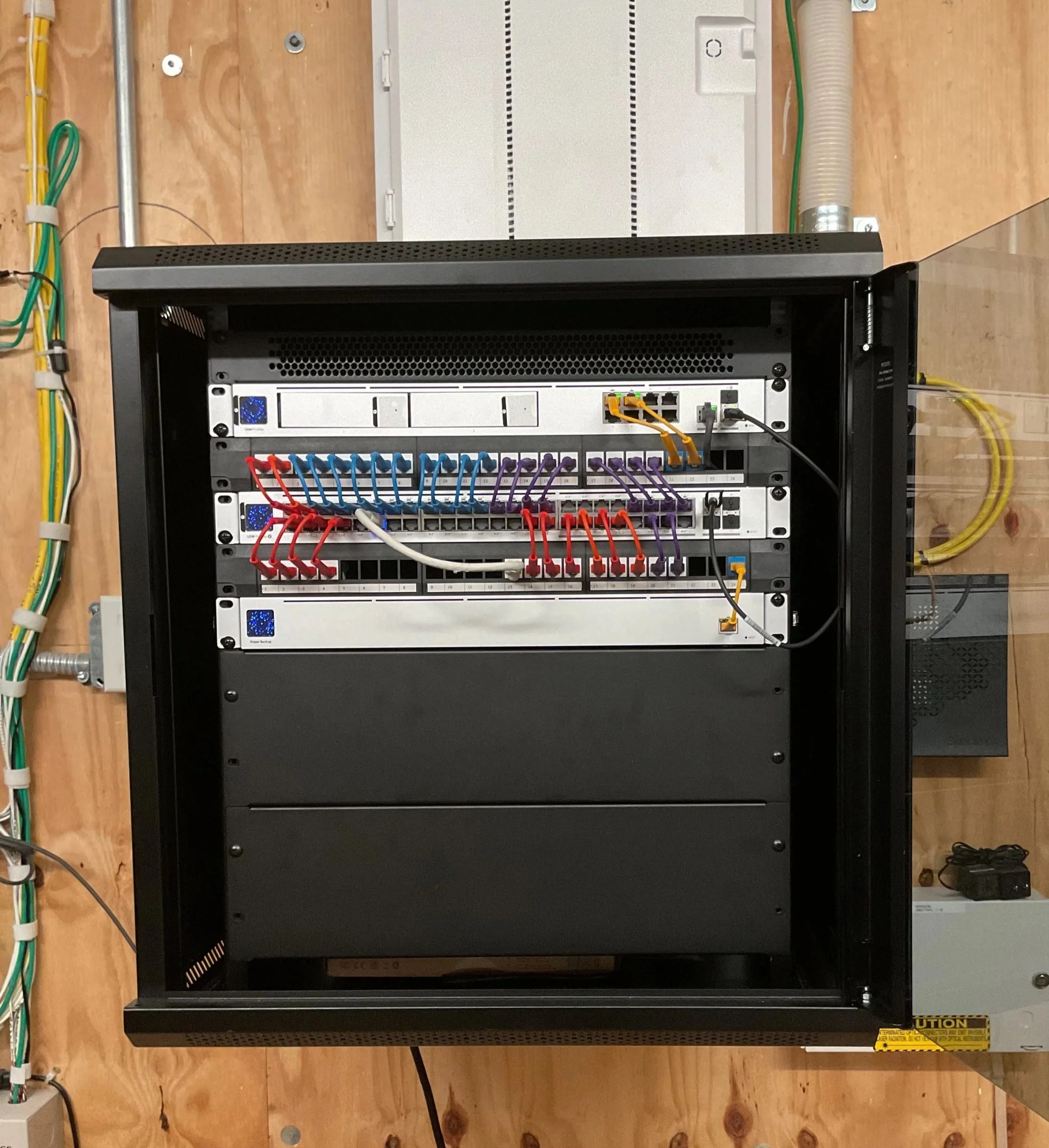 Open network equipment closet with various wires and devices installed on a wooden wall, including black server rack, patch panels, and visible Ethernet and fiber optic cables.