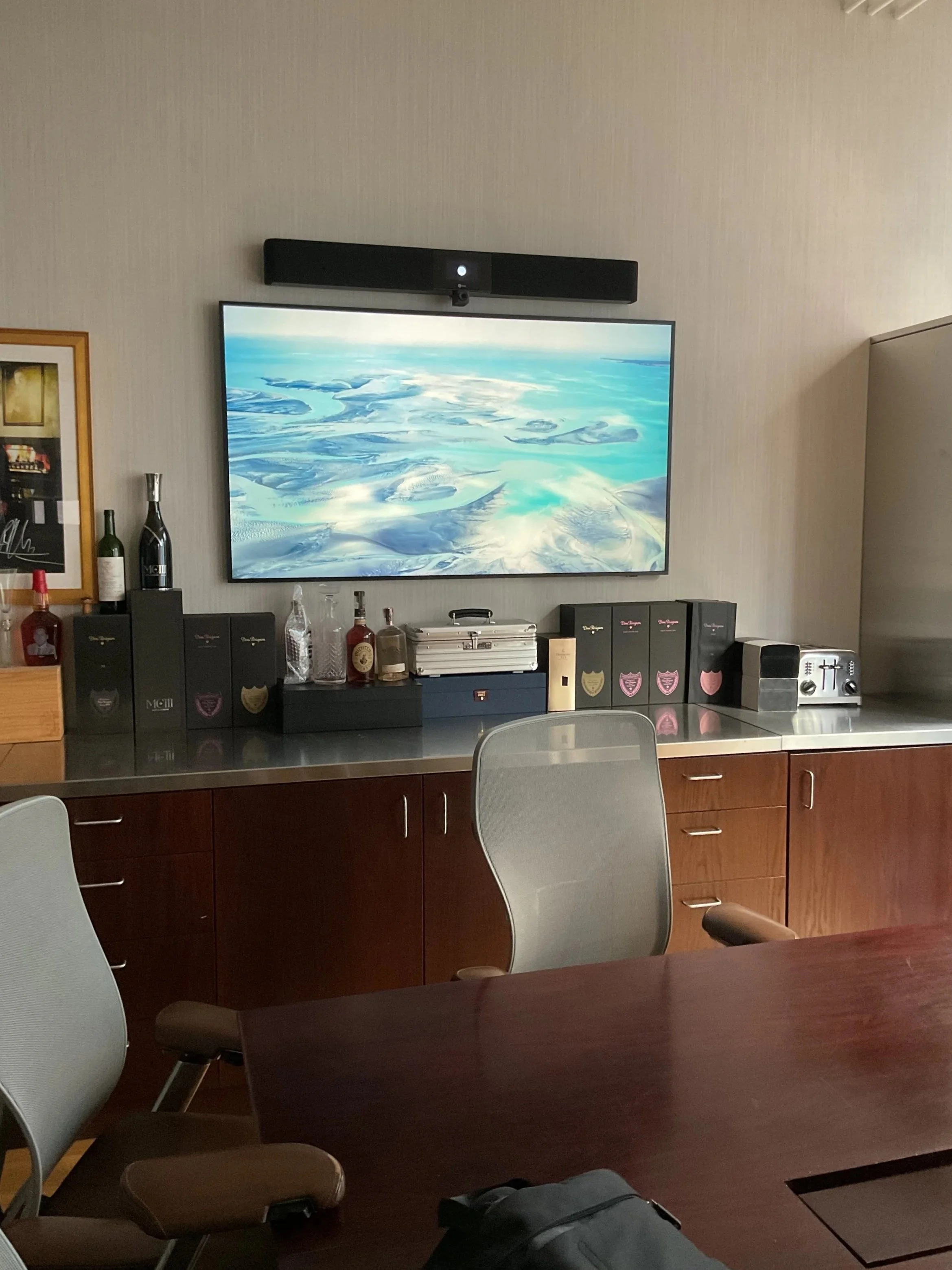 A flat-screen TV on a wall displaying an aerial view of icy blue water and snow-covered land. Below the TV, bottles and boxes are on a black surface, with a beige wall behind.