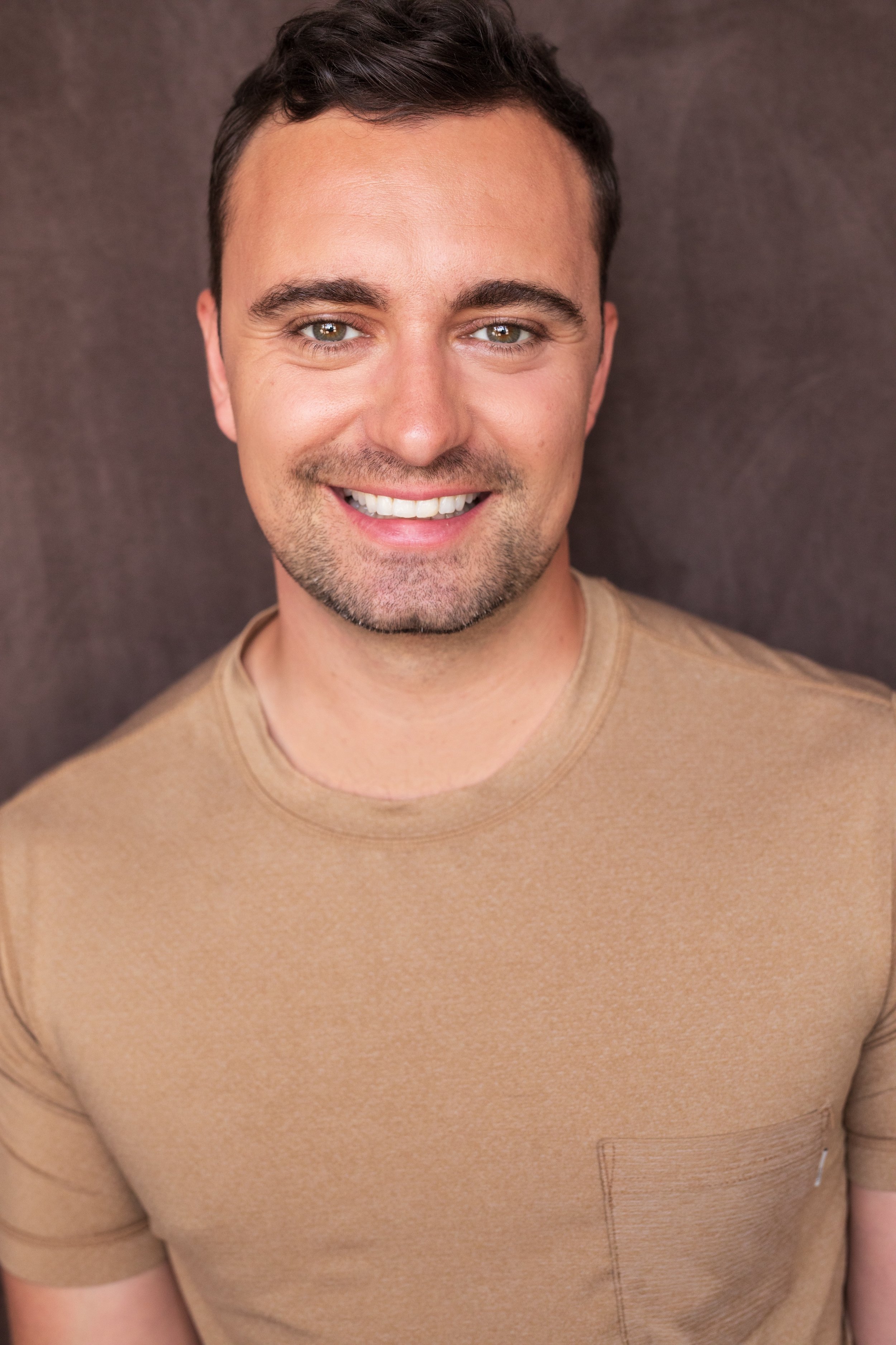 Close-up of a smiling man with short dark hair, light skin, and green eyes, wearing a tan T-shirt, against a dark background.