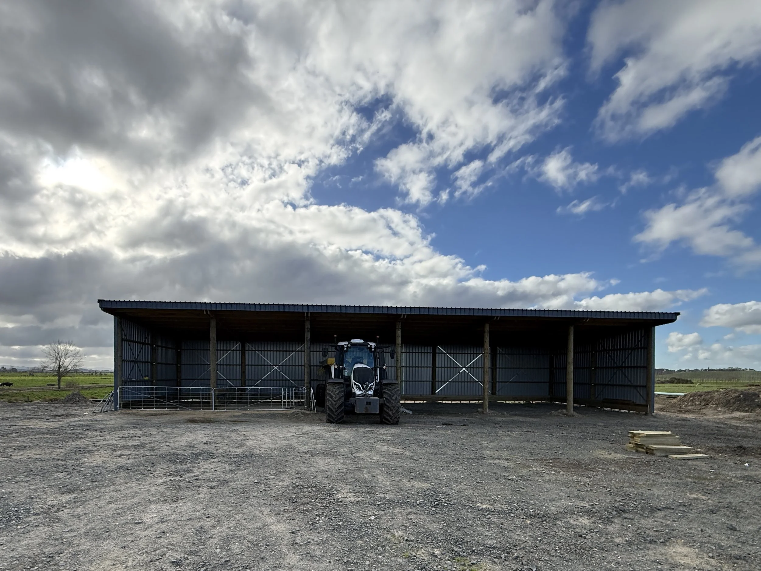 A farm building with a tractor parked inside, surrounded by open fields and a partly cloudy sky.