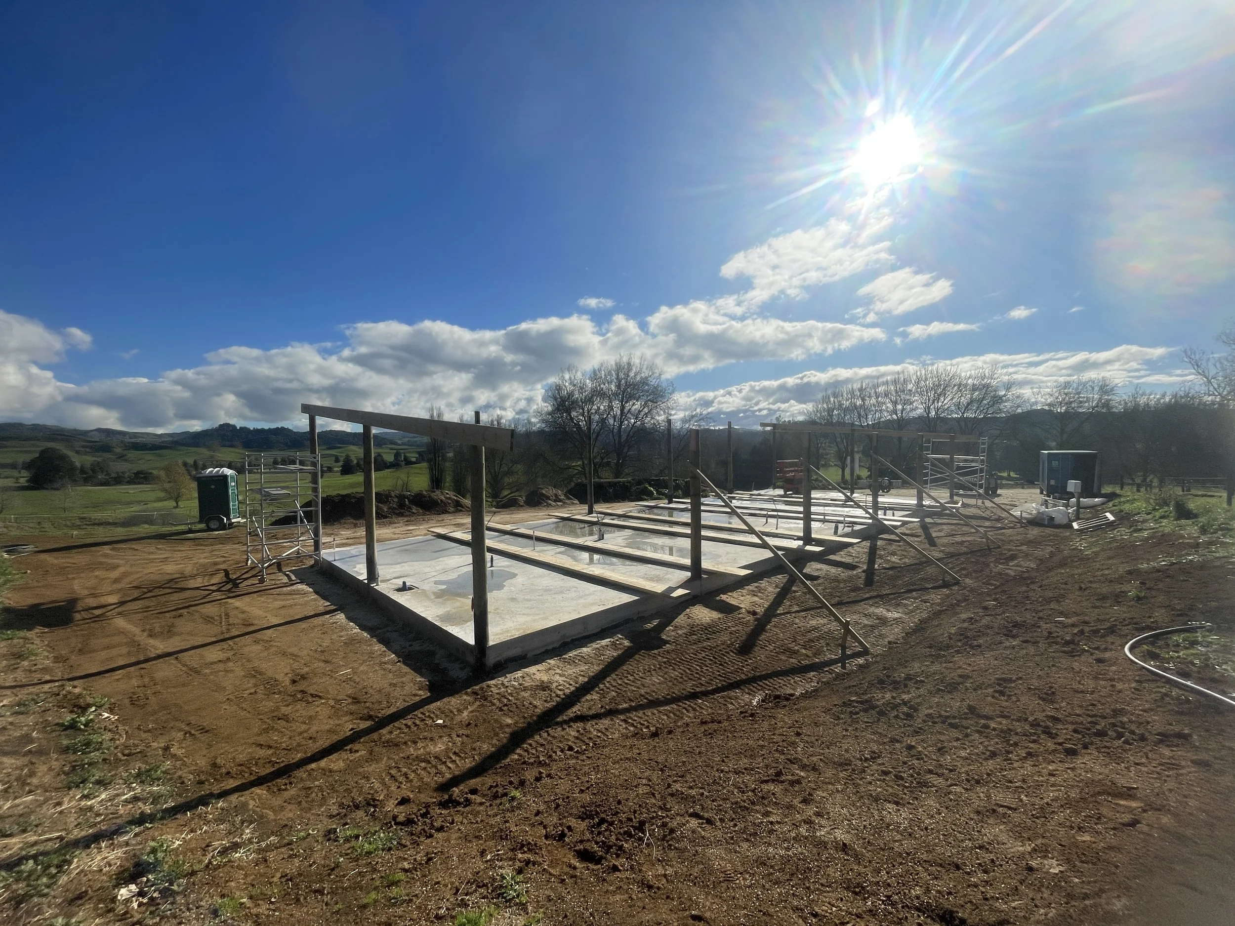 Construction site with building foundation, scaffolding, and construction equipment set against a bright sunny sky with scattered clouds and distant rolling hills.