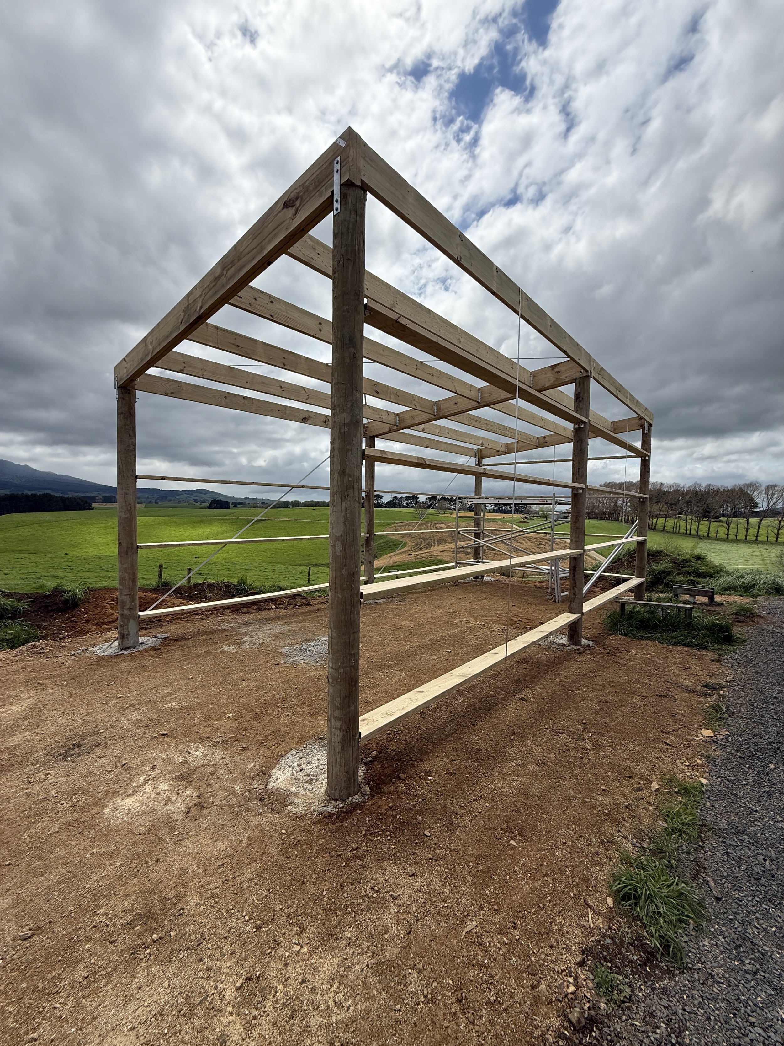 Wooden structure under construction on a dirt ground with green fields and a cloudy sky in the background.