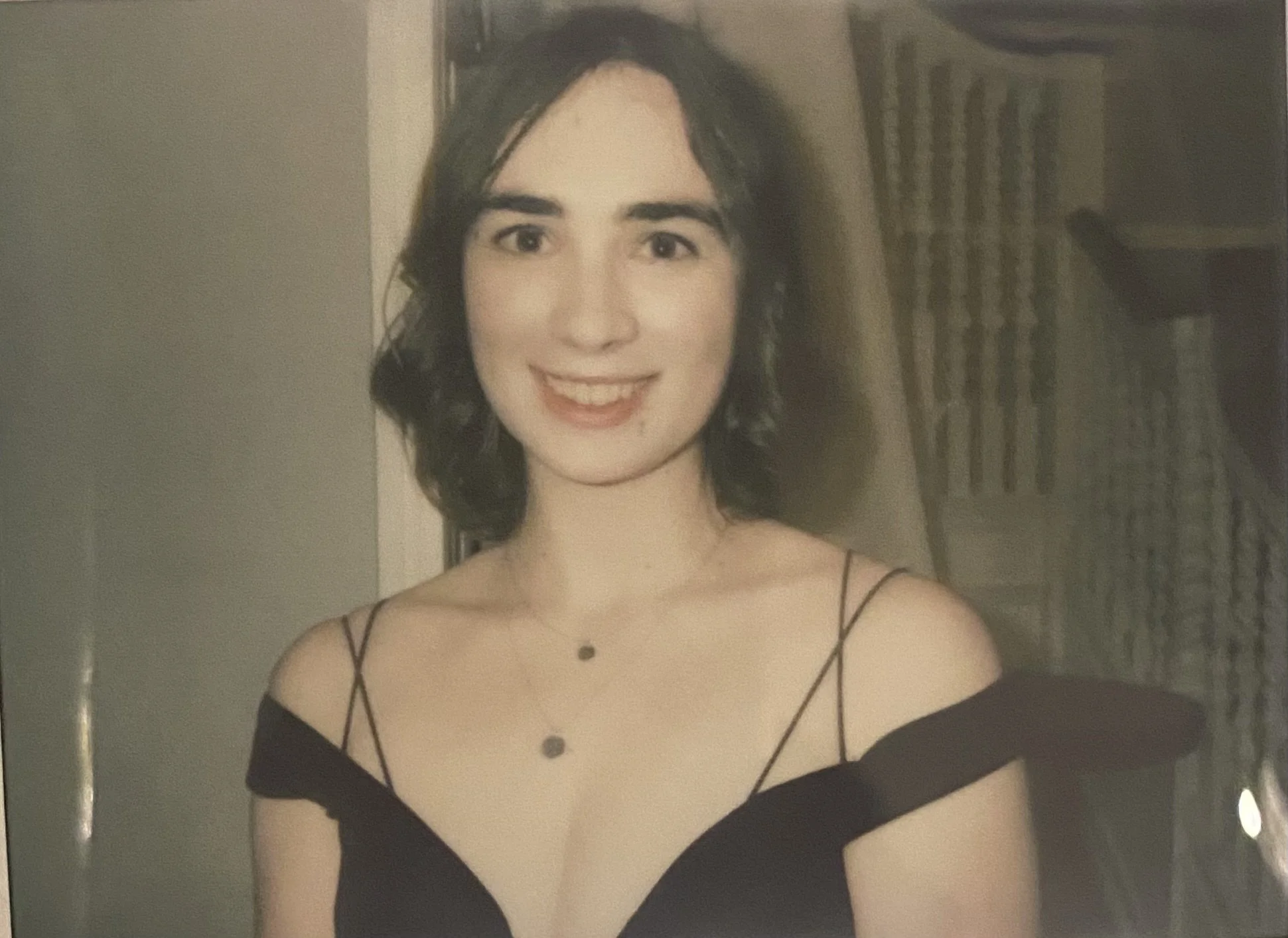 A woman with dark curly hair smiling, wearing a black off-shoulder dress with spaghetti straps and a necklace, standing in front of a plain wall with some furniture in the background.