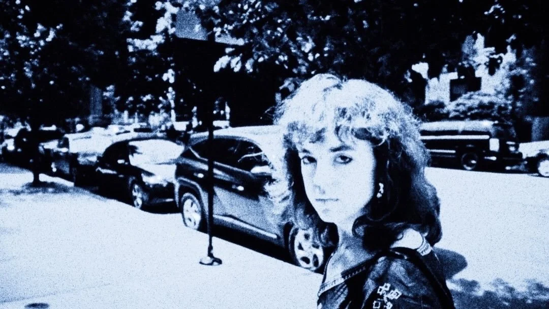 A woman with curly hair and earrings standing outdoors in a parking lot with parked cars and trees.