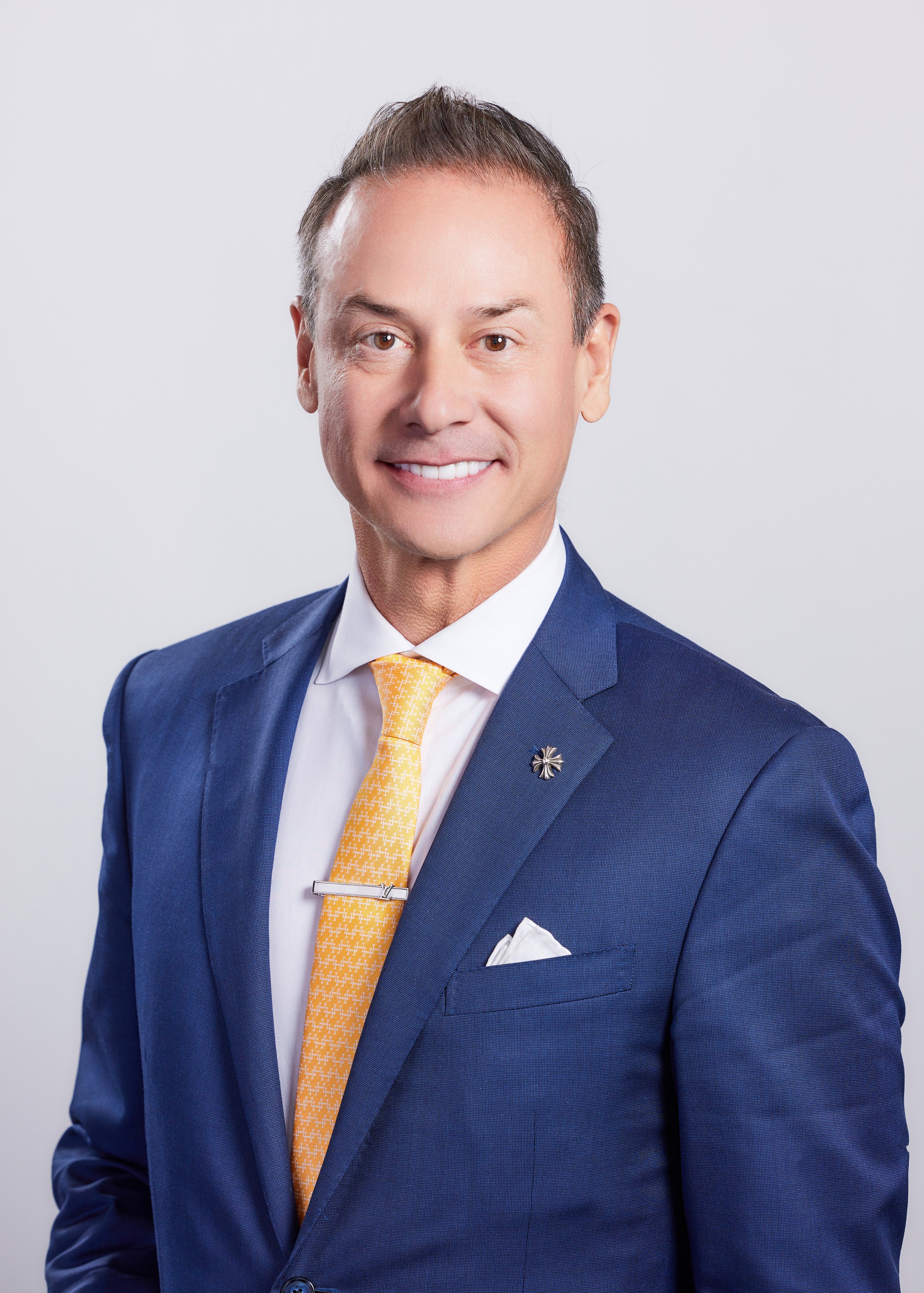 A man wearing a navy blue suit, white dress shirt, yellow patterned tie, and a lapel pin, smiling at the camera with a light gray background.