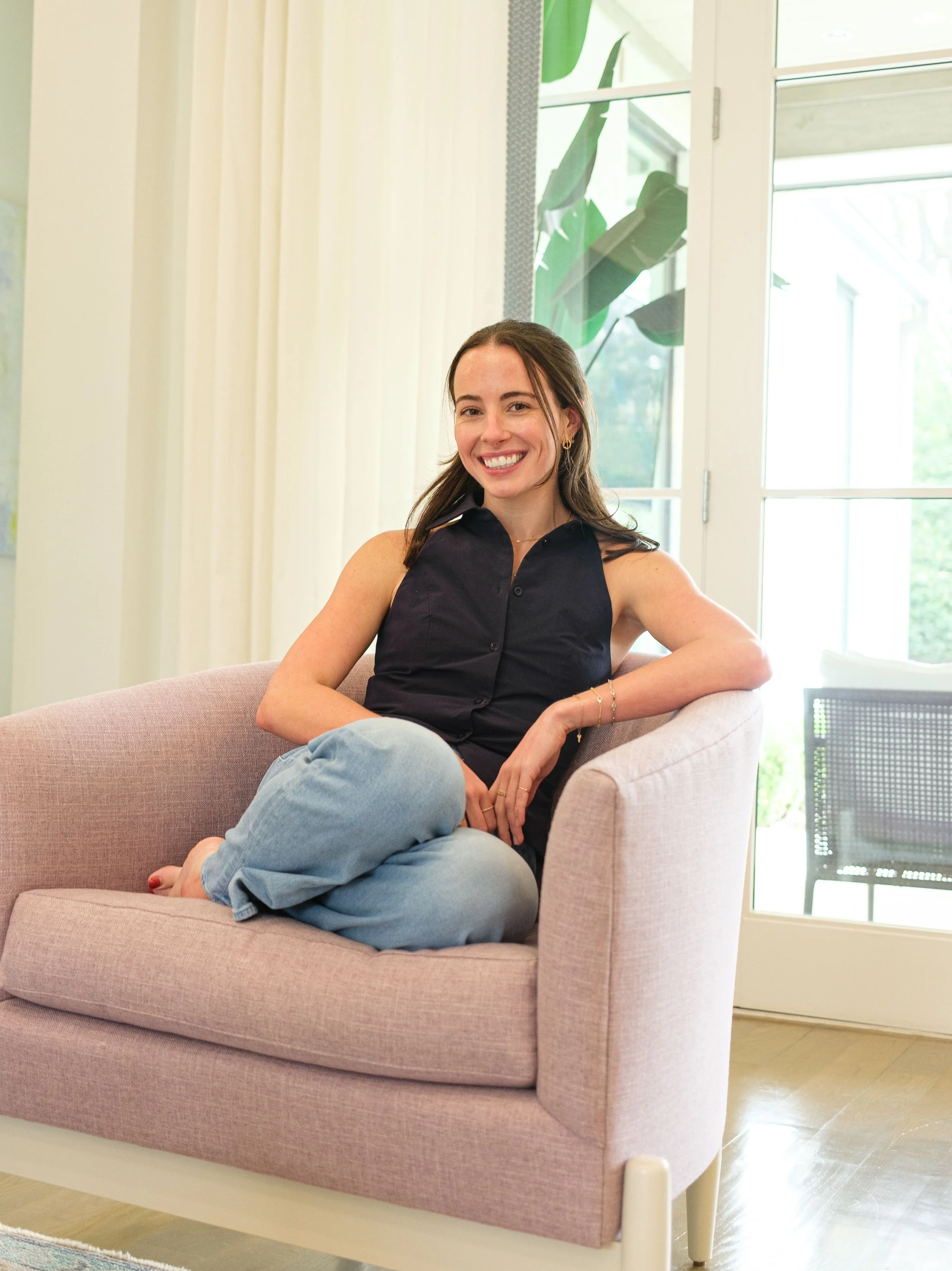A young woman with long brown hair, smiling, sitting on a pink armchair in a bright room near large windows, wearing a dark sleeveless top and light blue jeans.