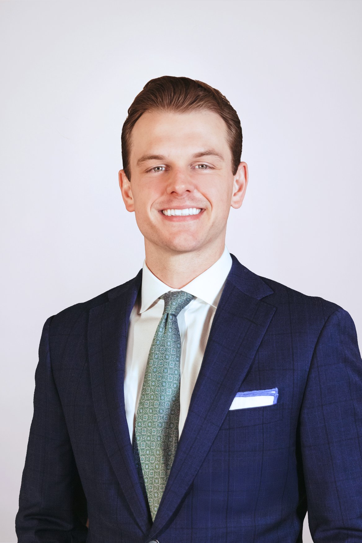 A young man with brown hair in a professional suit and tie, smiling, standing against a plain light background.