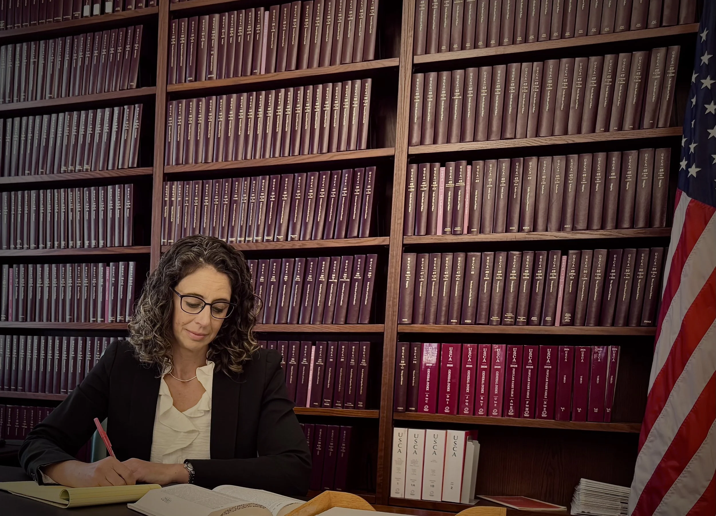 A woman with curly hair, glasses, and a black blazer is sitting at a desk, signing a document with a red pen, in front of a large wooden bookshelf filled with purple and red binders. An American flag is partially visible to the right.
