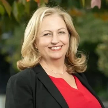 A woman with blonde hair smiling outdoors, wearing a black blazer and red top, with a blurred green background.