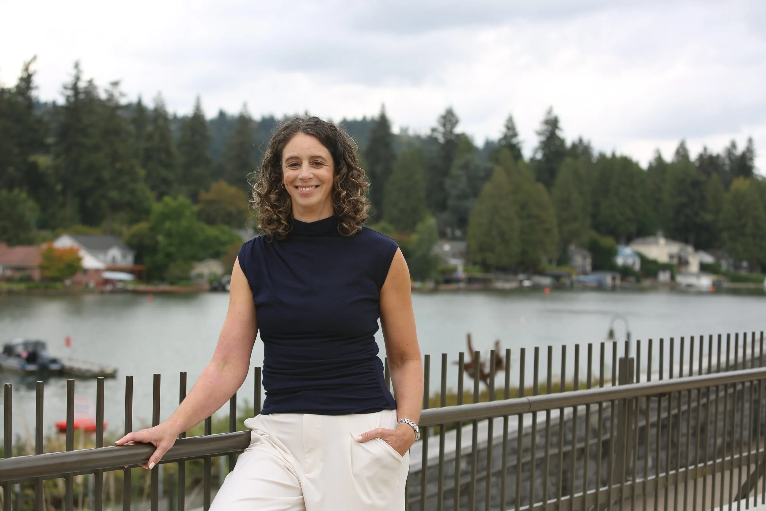 A woman with curly brown hair, wearing a black sleeveless top and white pants, stands outdoors by a metal railing near a body of water, with trees and houses in the background under a cloudy sky.