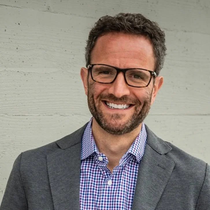 A man with curly dark hair, glasses, and a beard, smiling while wearing a blue checkered shirt and a gray blazer, standing in front of a light gray wooden wall.