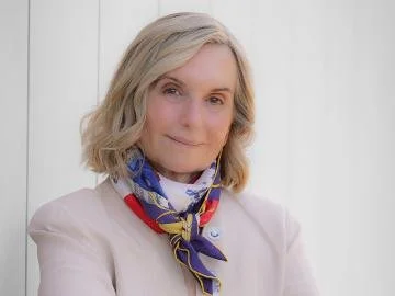 A woman with blonde hair, wearing a white blazer and a colorful scarf, standing against a white background.