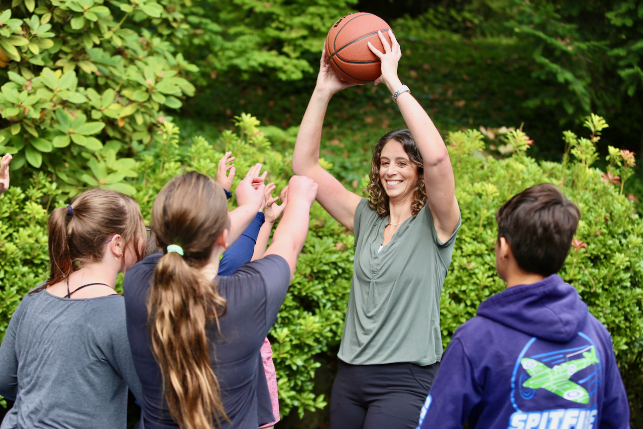 A woman smiling while holding a basketball overhead, surrounded by children reaching for the ball outdoors.