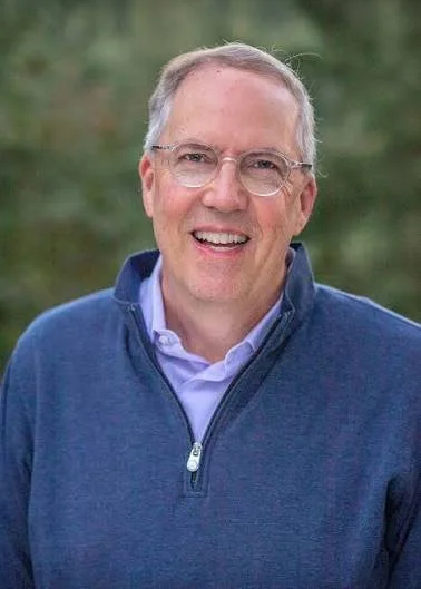 A middle-aged man with gray hair and glasses smiling outdoors, wearing a blue jacket and a light purple shirt, with a blurred green background.