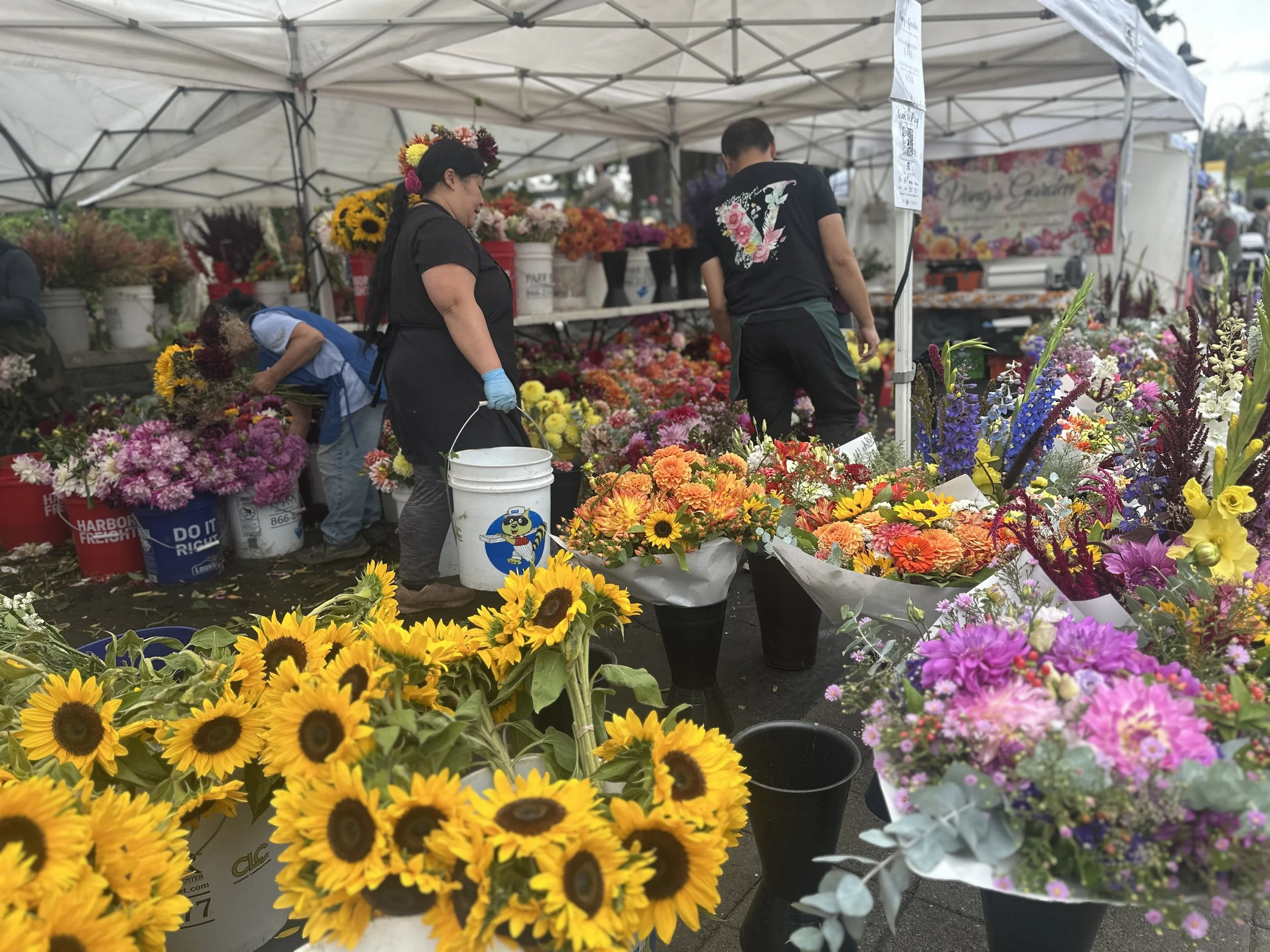 A flower market stall with colorful flowers including sunflowers, dahlias, and other bright blooms under a white canopy. People are shopping and arranging flowers.