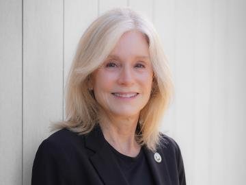 Portrait of a woman with shoulder-length blonde hair, wearing a black blazer, smiling, standing in front of a white wall.